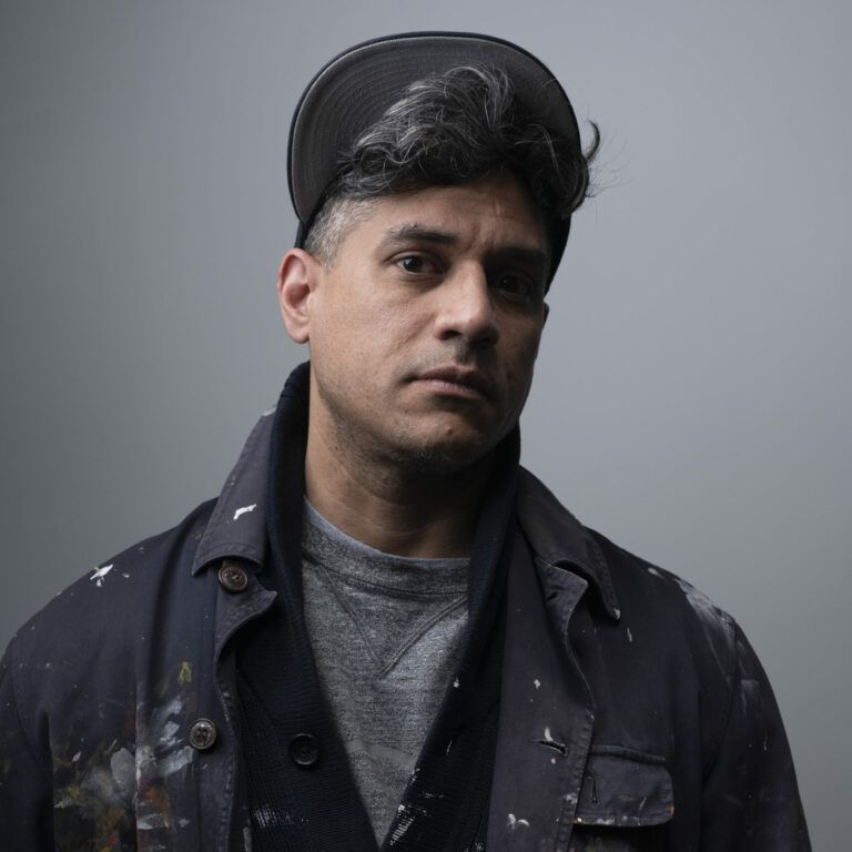 Color portrait of Josue Pellot. He wears a dark jacket and a dark baseball cap, standing in front of a grey background.
