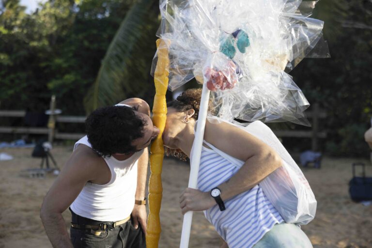 Two people licking mango limbers stacked on the same pole. The iced treat melts in the heat of the public outdoor area.