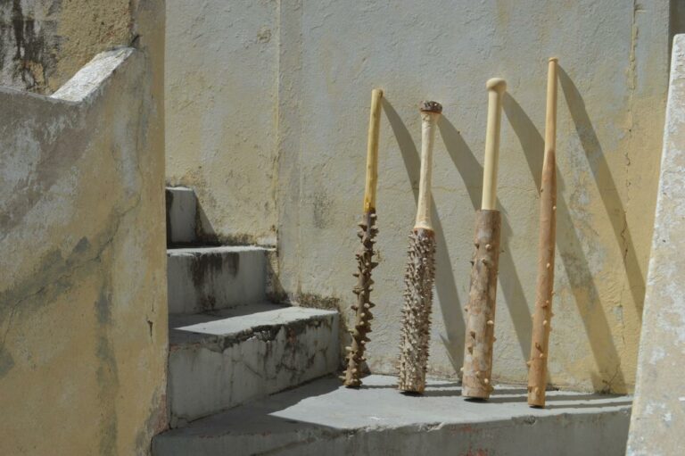 Four wooden bats carved from sharp thorny logs. They are leaned up against a beige concrete wall.