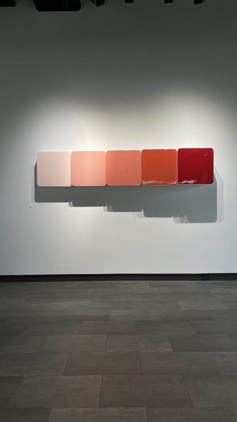 Five reflective resin squares mounted on a white wall. Each rounded square is a slightly more tinted red color from left to right.