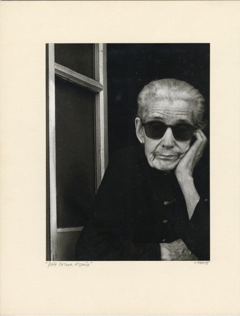 Portrait of an older person in a windowsil wearing sunglasses and resting their head in their hand.