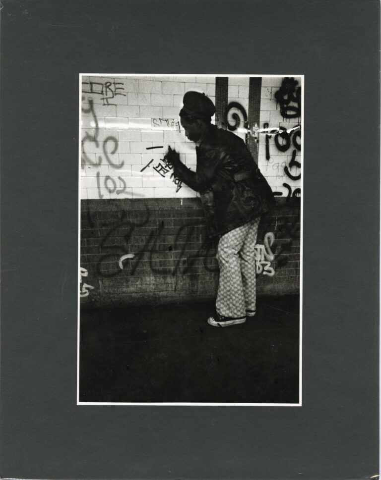 Photograph of a man writing on subway tile, his writing among tags left by others before him.