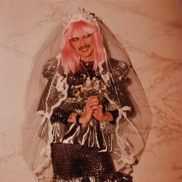 Color image of someone with a pink wig and a wedding veil holding flowers and smiling.