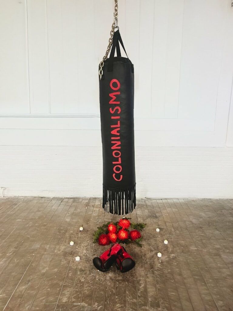 A black punching bag with the word "Colonialismo" painted on the side in red. Under the bag are red flowers and red boxing gloves.