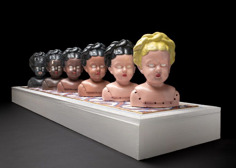 six doll heads are cast in porcelain and arranged in a line from light skin to dark skin, with the lightest doll facing first and forward and each subsequent doll head facing behind her.