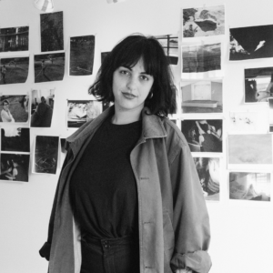 Black and white photo of artist Genesis Baez in front of a wall covered in photographs.